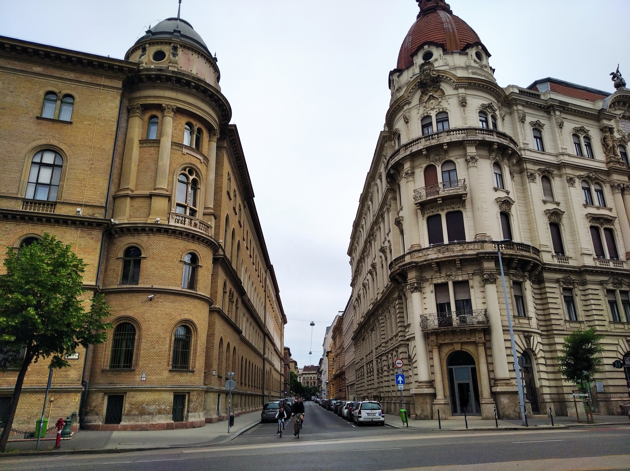 A pair of grand Belle Époque buildings on a quiet Buda street, with a single cyclist passing by