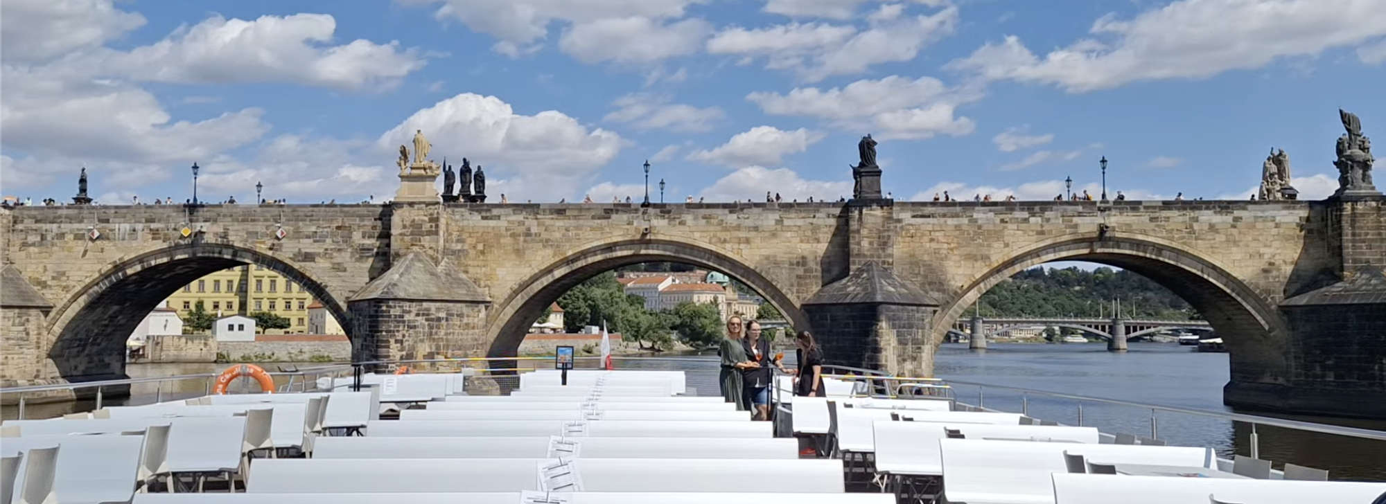 Wide panoramic view of Charles Bridge arches from a sightseeing boat passing underneath, white deck chairs in the foreground