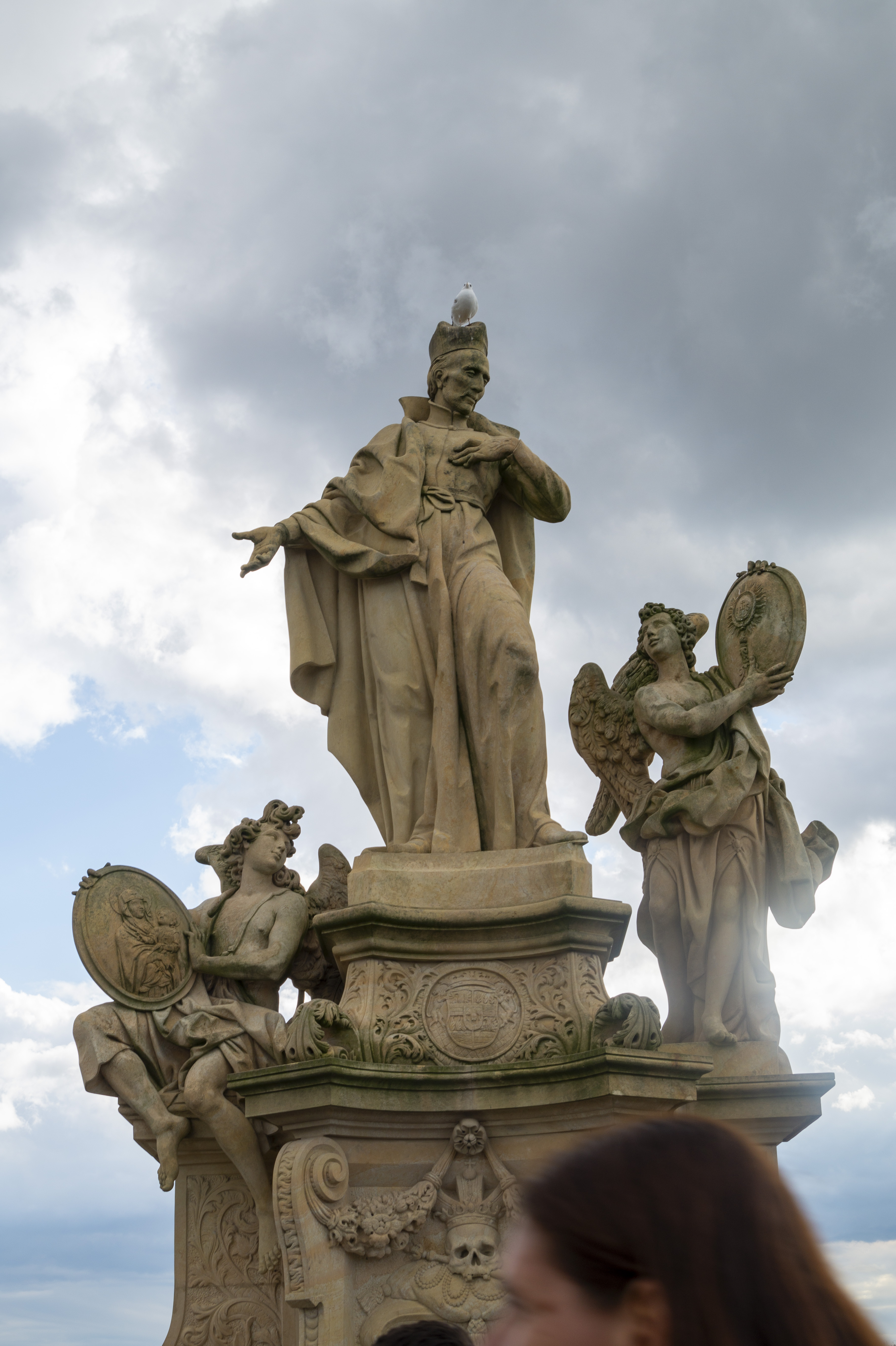 Baroque statue on Charles Bridge — a saint in long robes flanked by two angels with mirrors, dramatic clouds behind, a seagull perched on the saint's head