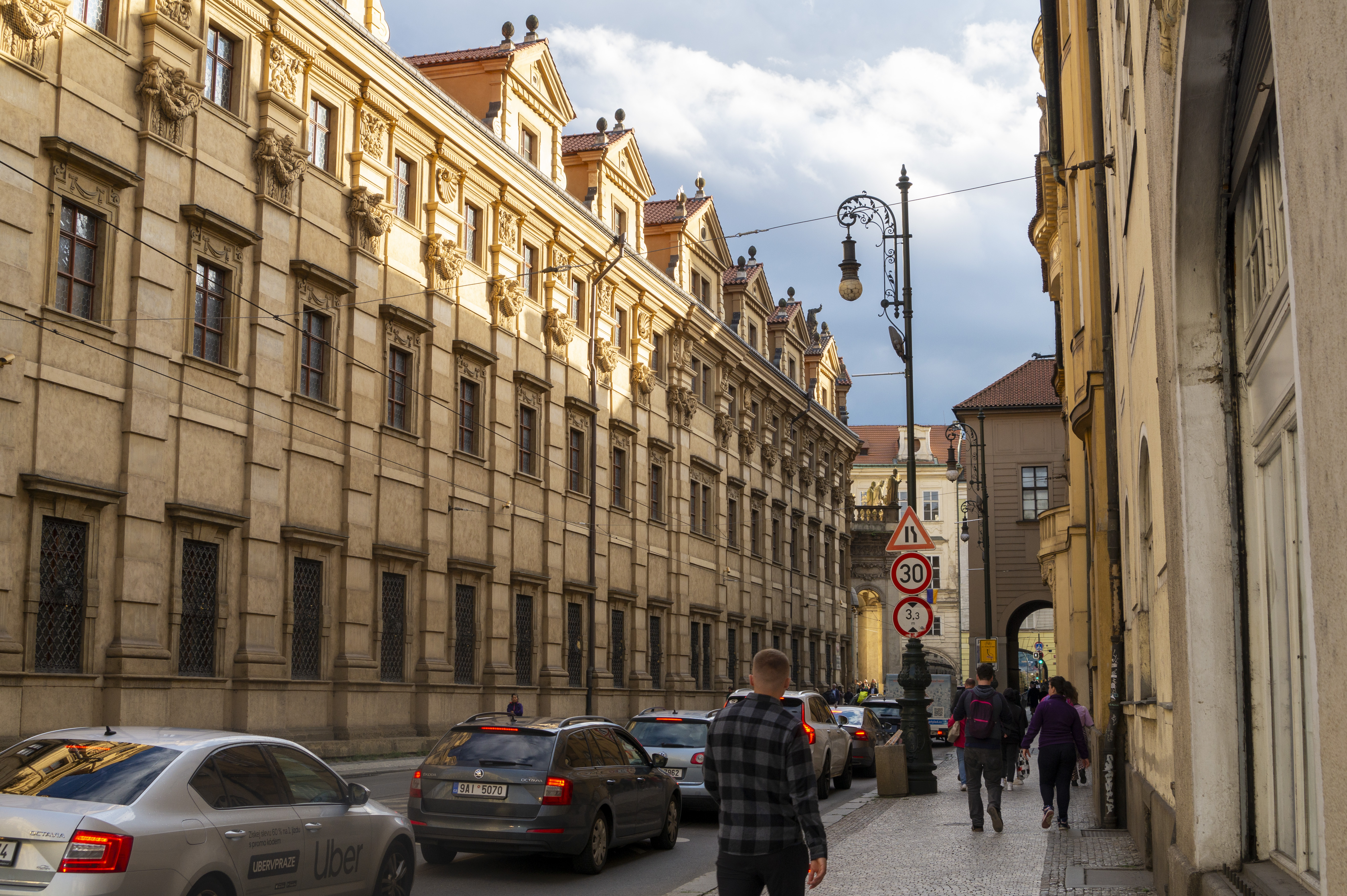 The honey-coloured baroque facade of Clam-Gallas Palace stretching down a narrow Old Town street with vintage lamp posts and afternoon golden light