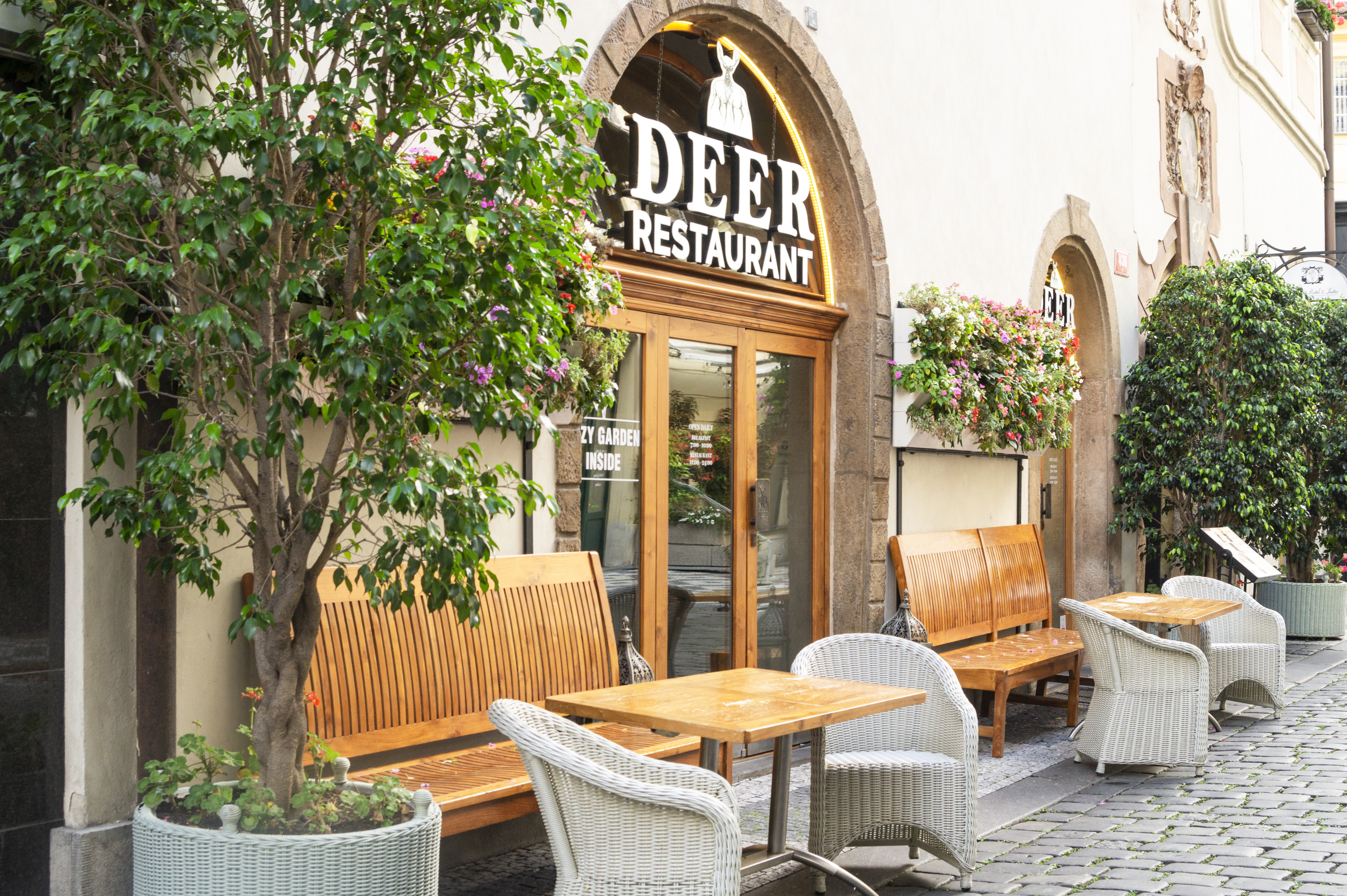 Outdoor terrace of DEER Restaurant in Prague with wooden chairs and tables under a stone archway entrance, flowering bushes by the door