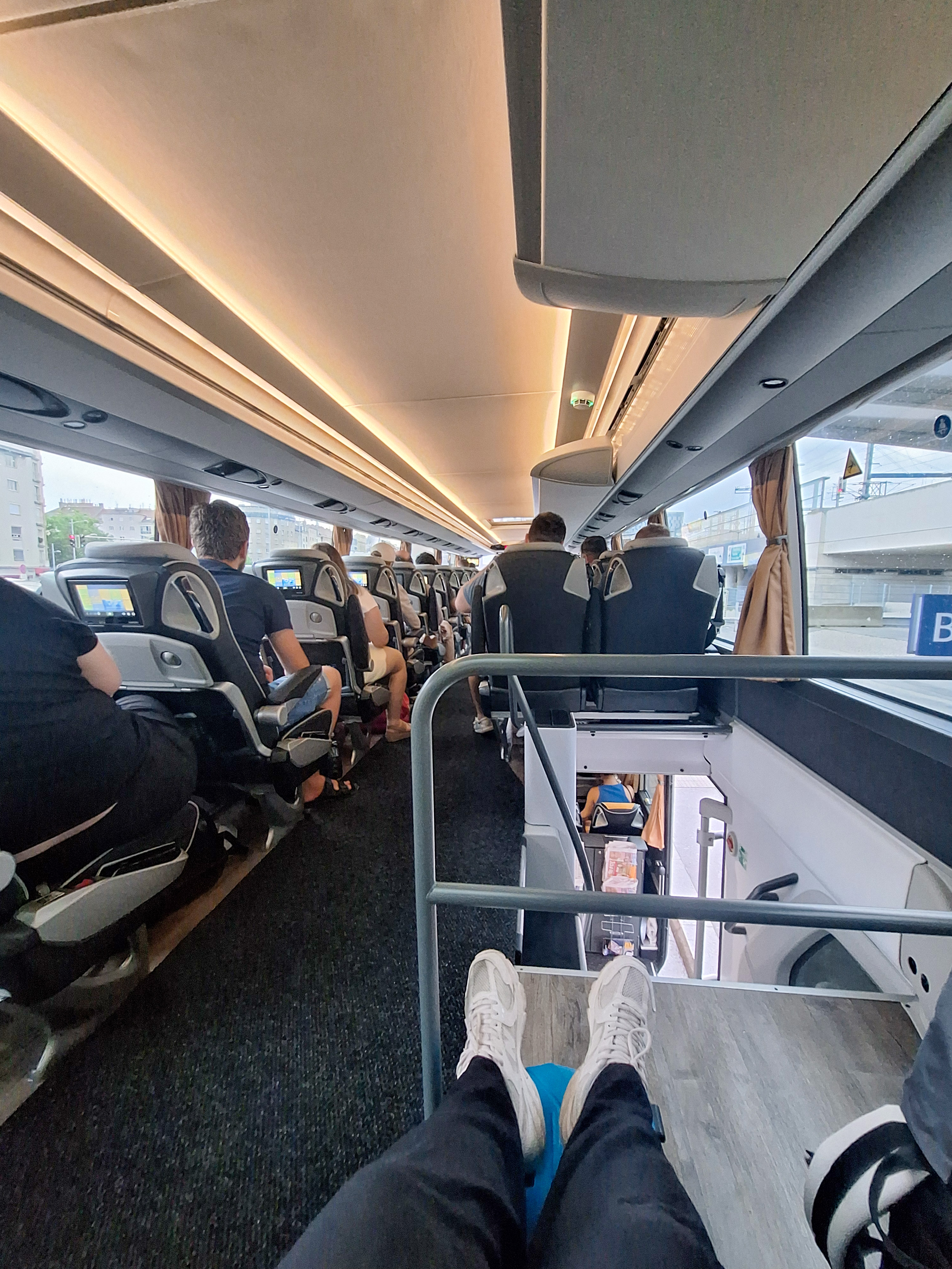 View down the upper deck of a Flixbus double-decker bus from the front seat, passengers in rows below, morning sunlight coming through the panoramic windscreen