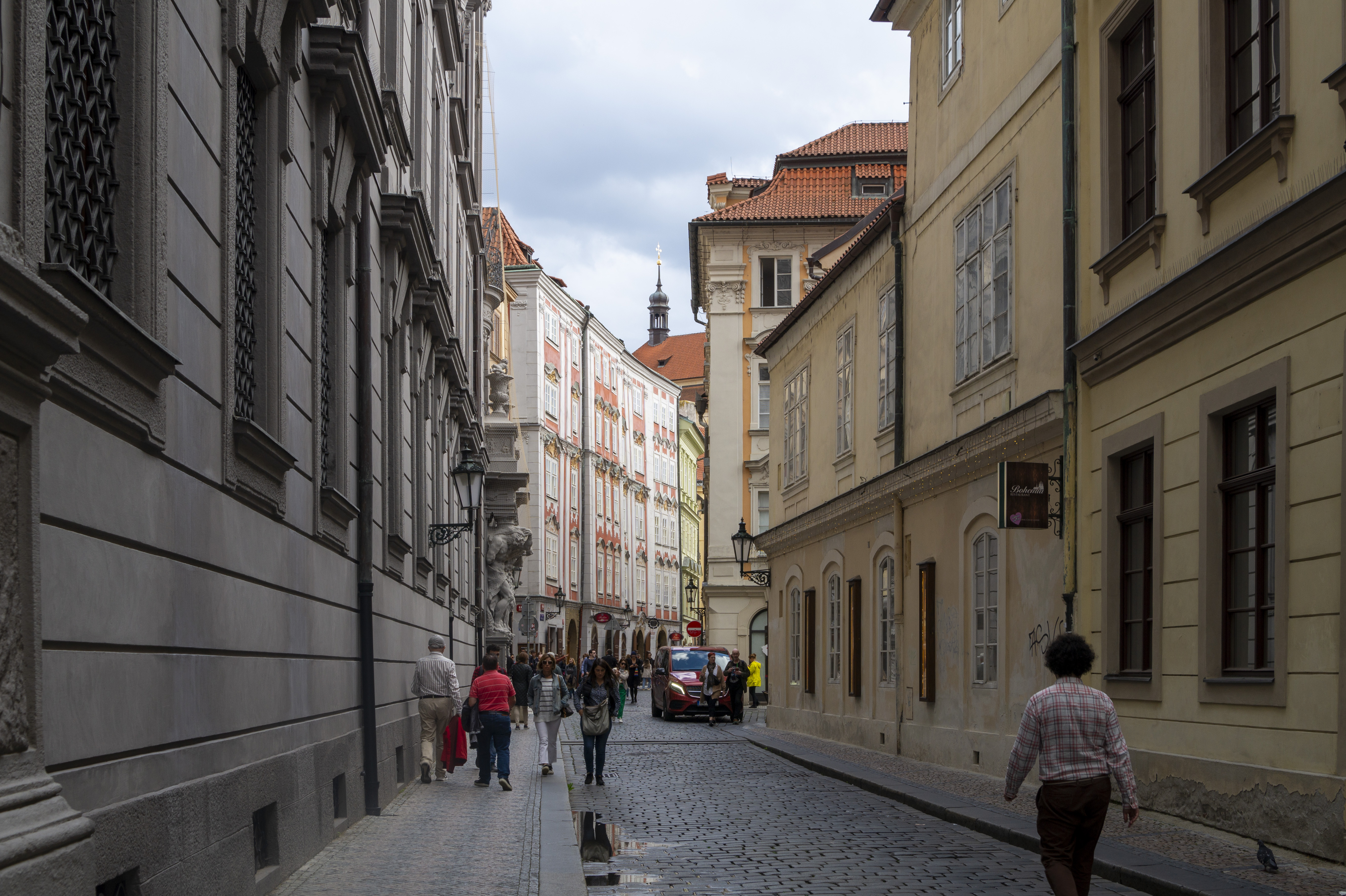 A narrow cobblestone side street in Prague's Old Town with rows of pastel baroque buildings, locals walking past, soft grey afternoon light