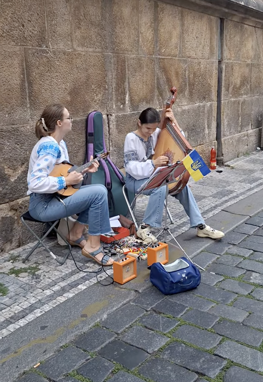 Two Ukrainian young women in embroidered vyshyvanky shirts playing bandura and lute on a Prague street, Ukrainian flag draped on the bandura case beside them