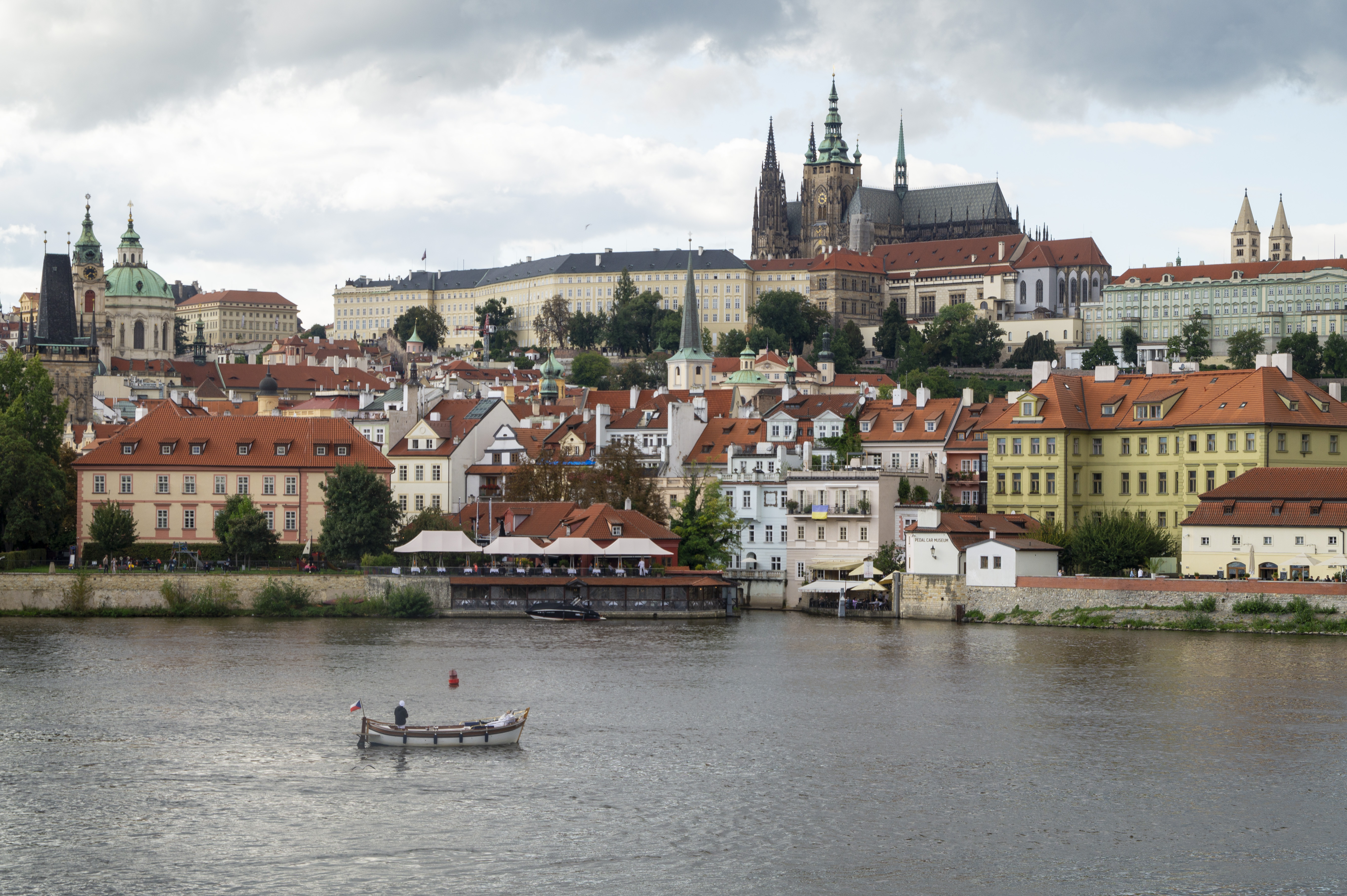 View across the Vltava to Prague Castle and St Vitus Cathedral on the hill, with red-tiled rooftops of Malá Strana below and a small wooden boat on the river