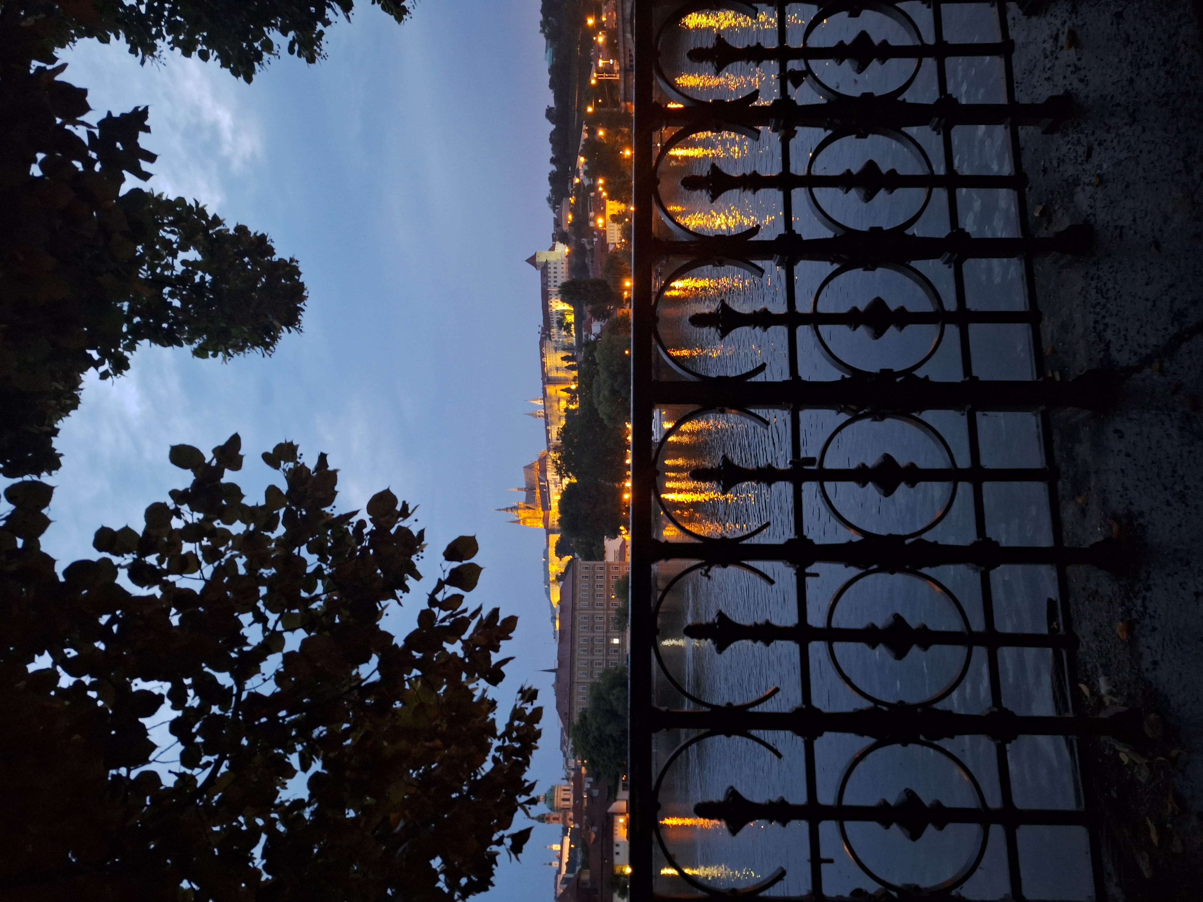 Evening view of the Vltava embankment in Prague at twilight with golden street lamps reflected in the calm water, an ornate iron railing in the foreground and Prague Castle silhouetted on the horizon