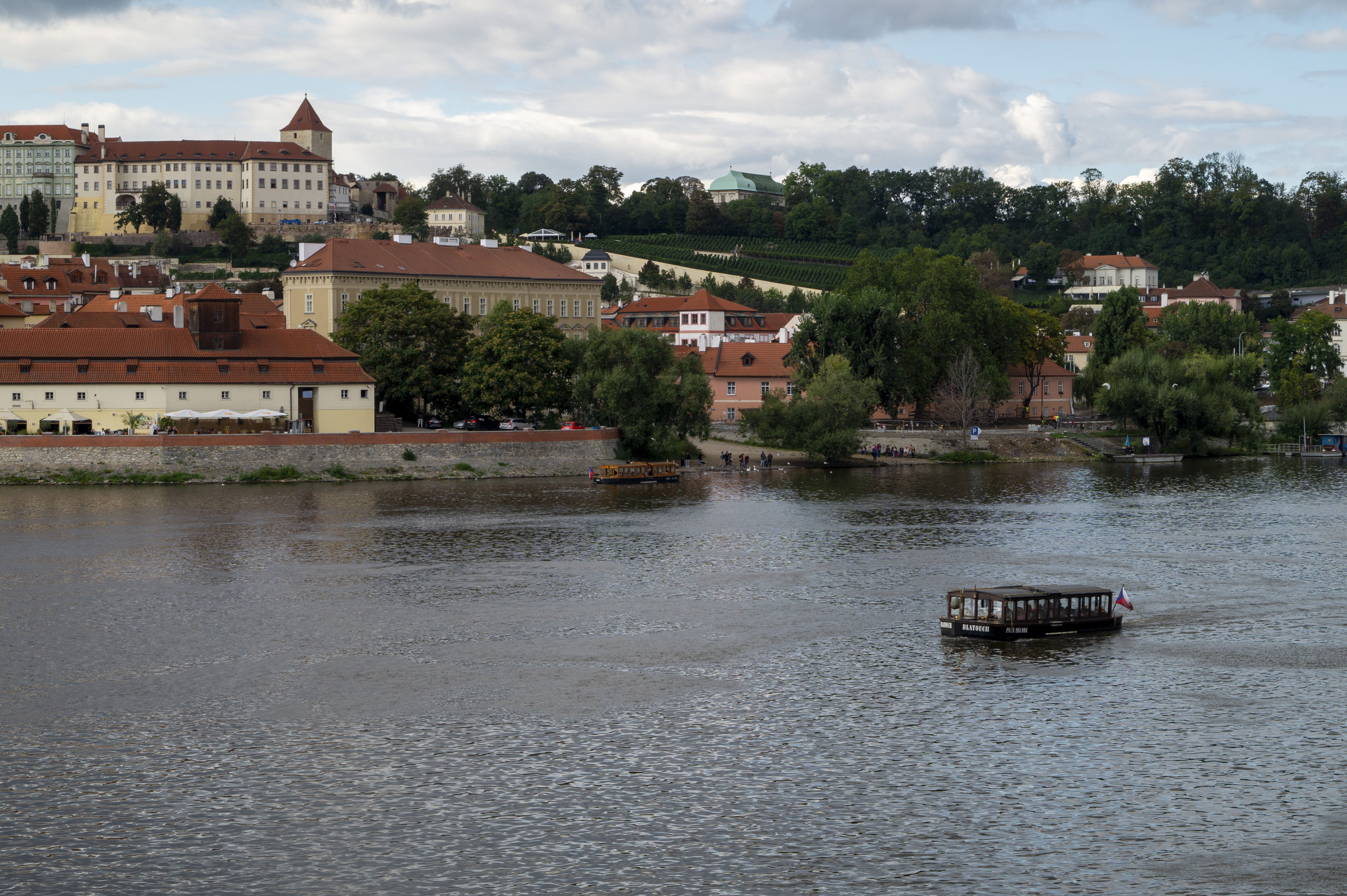 View across the Vltava to the white renaissance Royal Summer Palace and the green slopes of Letná Hill, with a small wooden ferry boat crossing the river