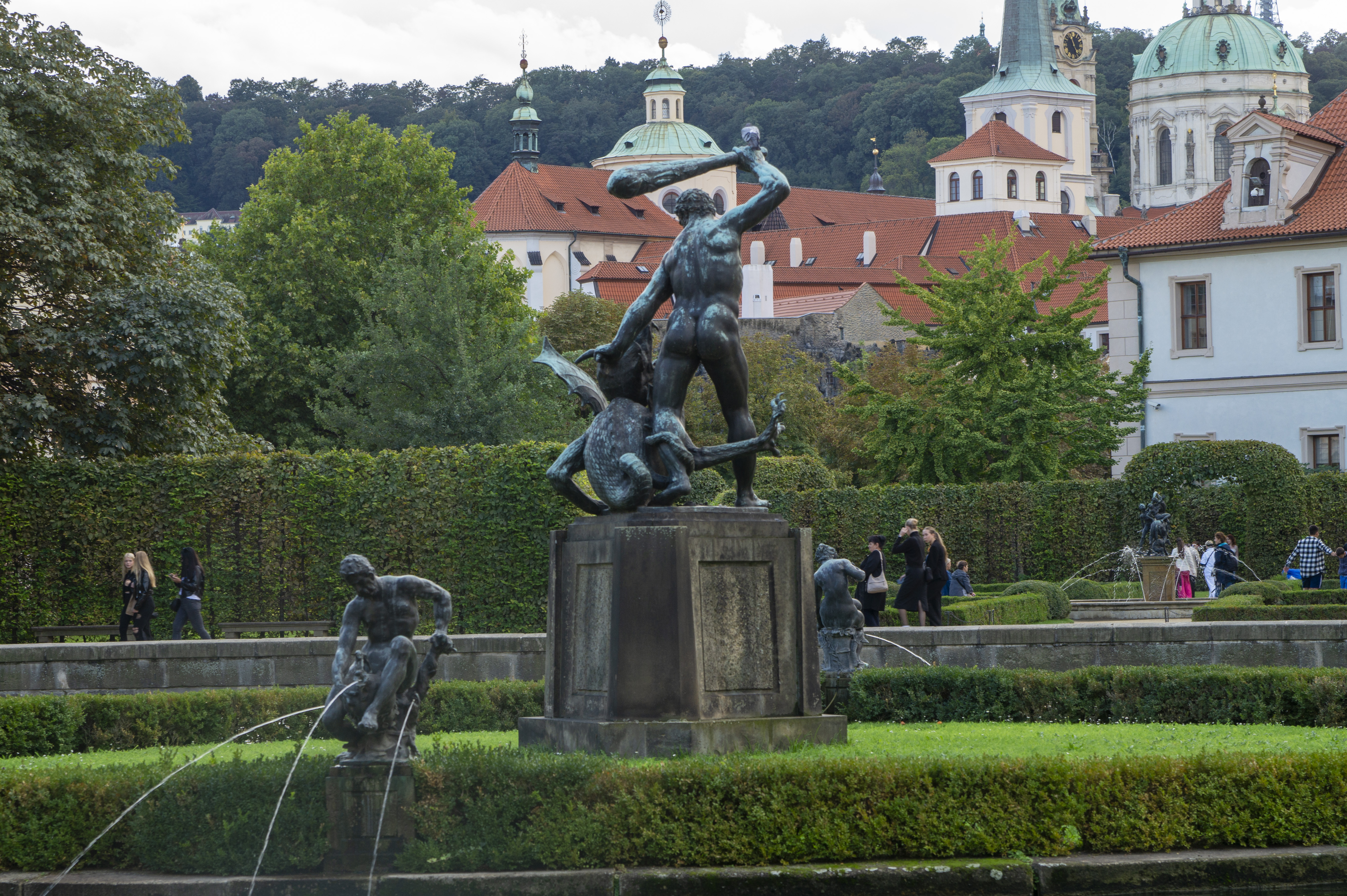 Bronze fountain statue of Hercules wrestling a lion in Wallenstein Garden in Prague, with manicured hedges, the green domes of St Nicholas Church and red-tiled rooftops behind