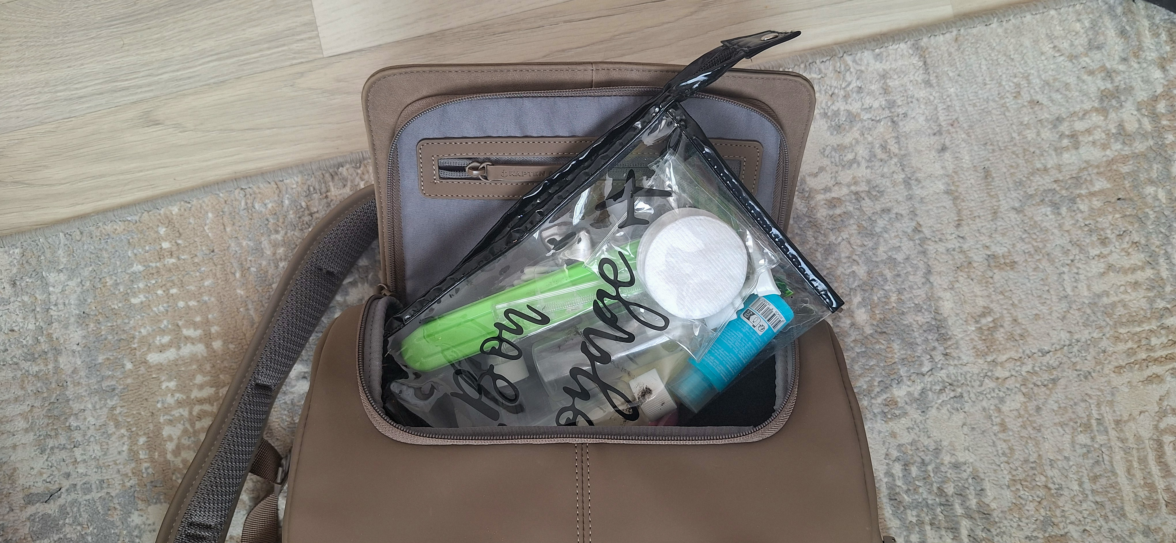 Transparent cosmetics pouch with travel-size bottles sitting on top of the open Kapten & Son backpack