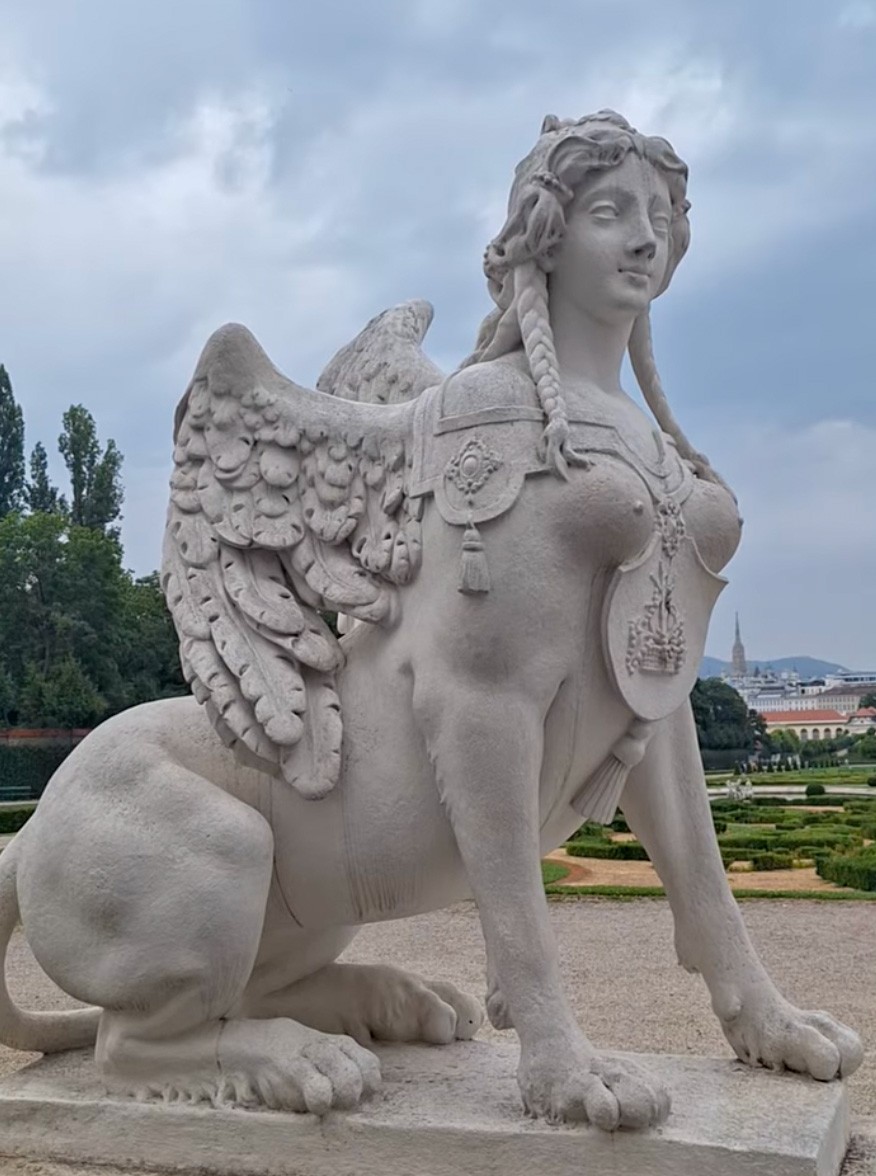 Baroque female sphinx statue in the Belvedere gardens — winged, with braided hair, looking out over the parterre toward the distant Stephansdom spire