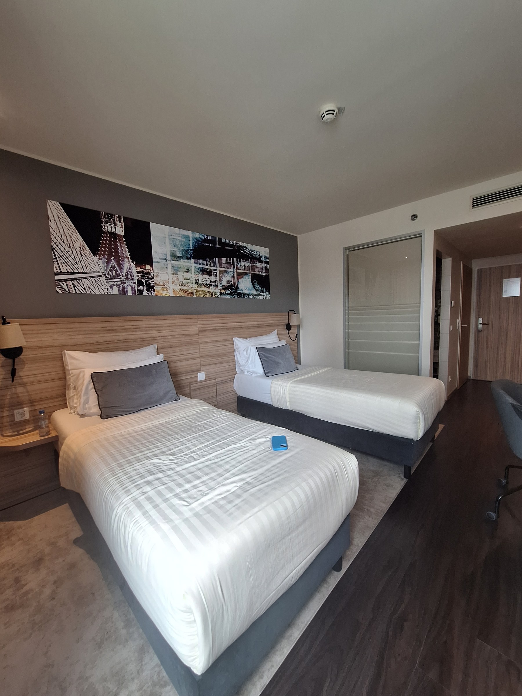 Twin room at Leonardo Hotel Vienna Hauptbahnhof — two beds, wooden headboard, grey wall and a framed black-and-white Stephansdom print above