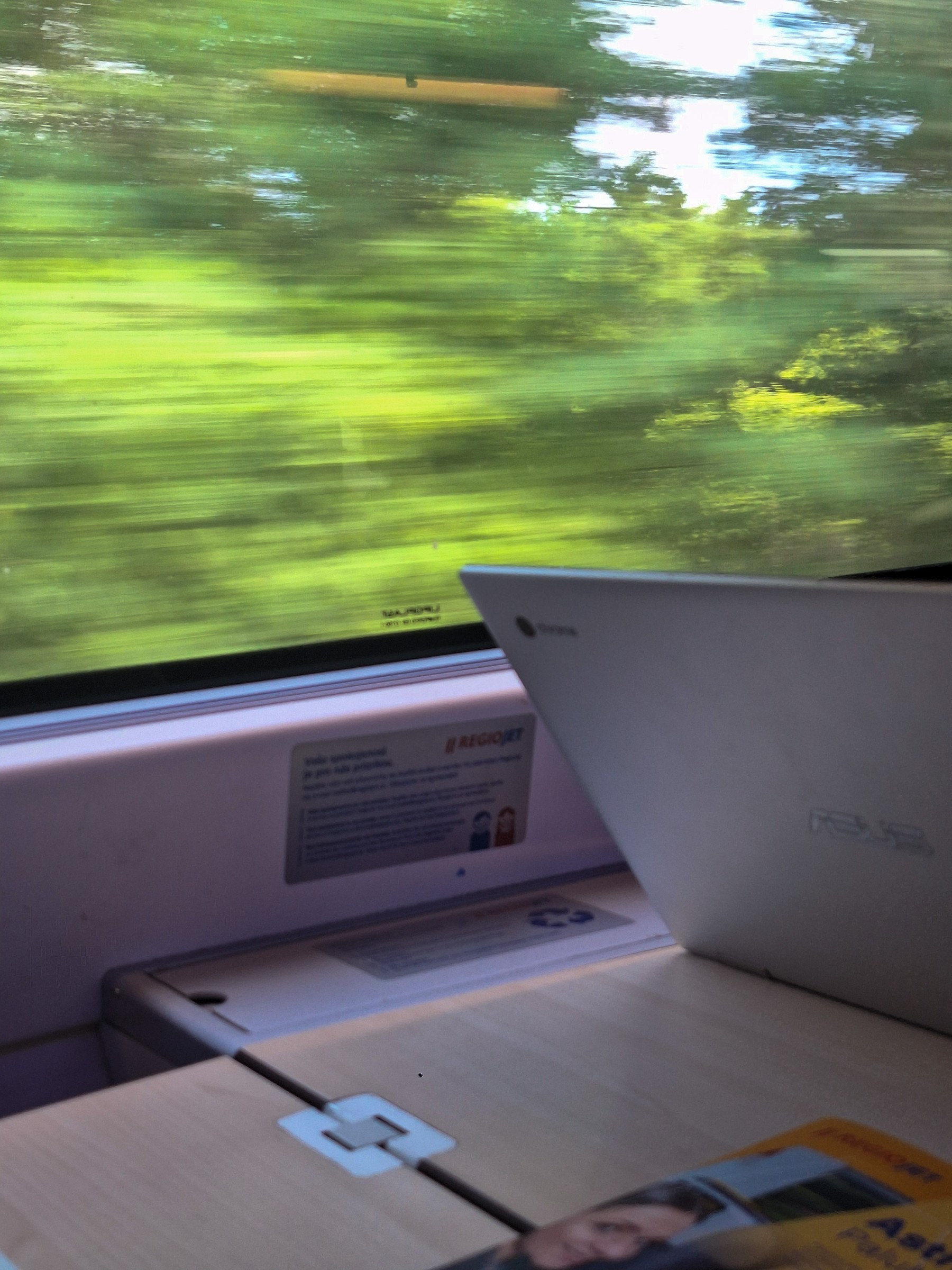 View from inside a RegioJet train window — motion-blurred green summer trees rushing past, laptop open on the tray table, RegioJet sticker on the window frame